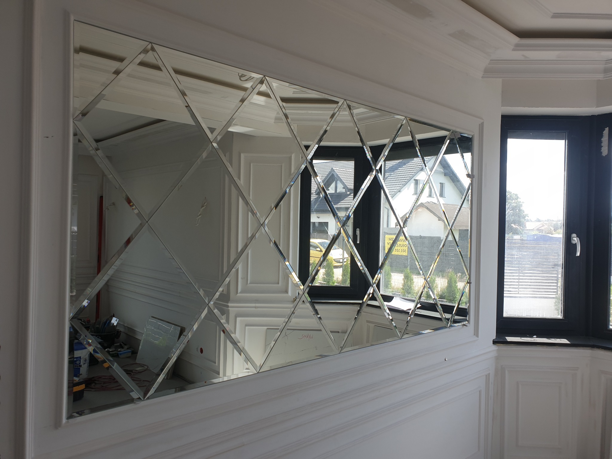 Dekoracyjne lustro ścienne z fazowanych rombów, odbijające wnętrze pokoju i widok z okna, zamontowane na białej ścianie z dekoracyjnymi panelami.