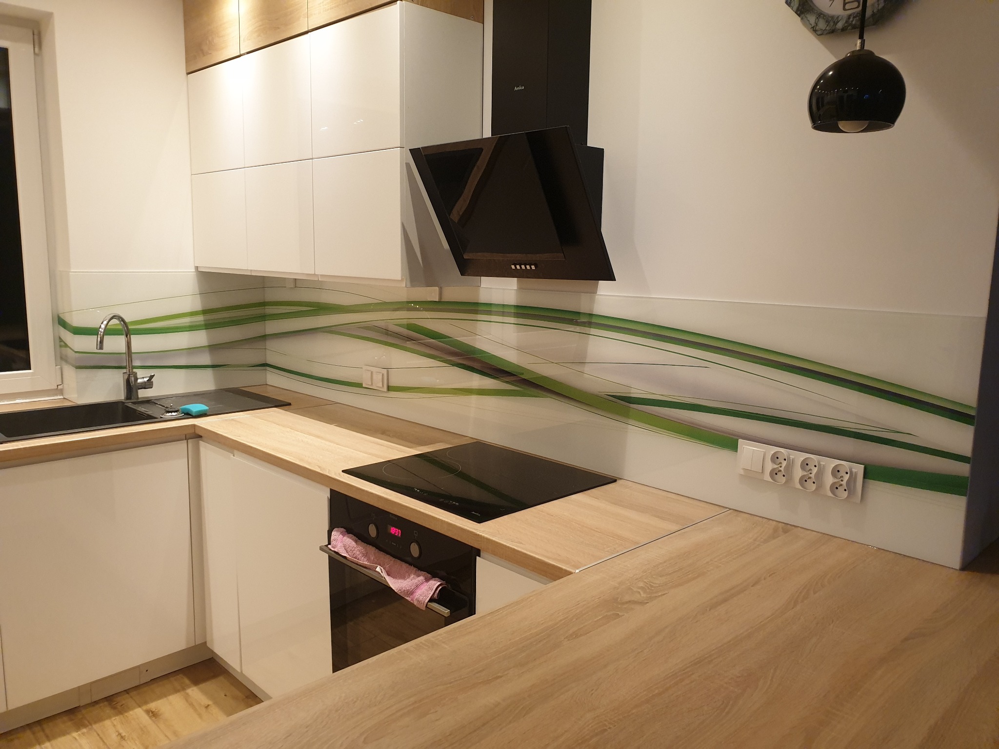 Nowoczesna kuchnia z białymi szafkami, drewnianym blatem i szklanym panelem ściennym z zielonym wzorem fal. Widoczny czarny zlewozmywak, płyta indukcyjna i okap.