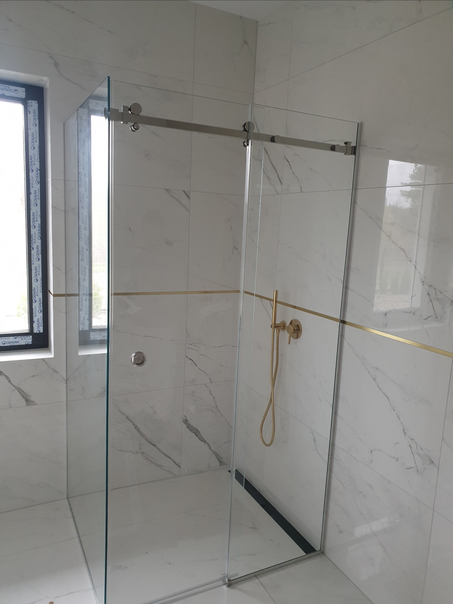 Szklana kabina prysznicowa z przesuwnymi drzwiami, chromowanymi okuciami i złotą armaturą na tle białych płytek imitujących marmur.