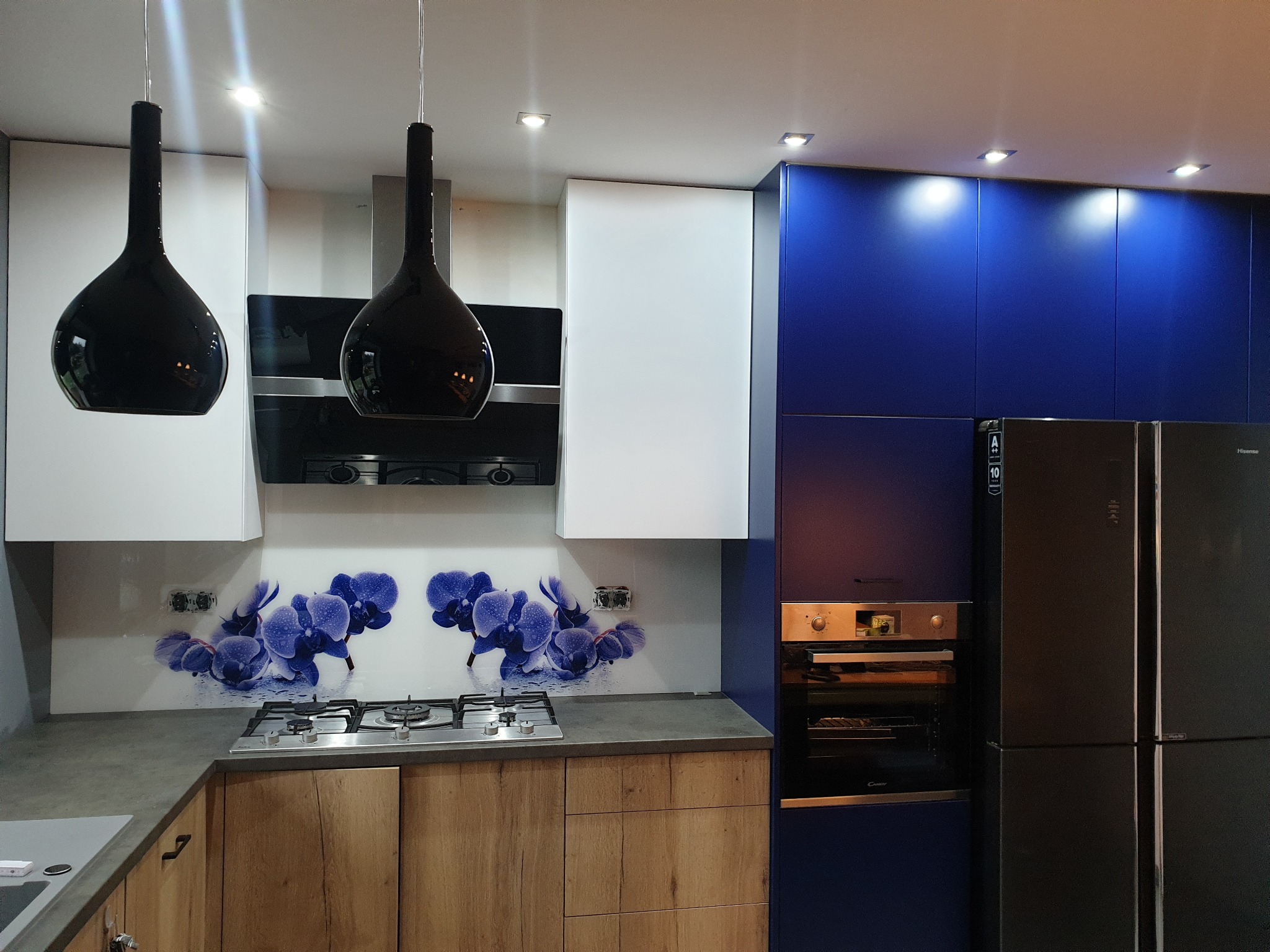 Nowoczesna kuchnia z drewnopodobnymi szafkami dolnymi, białymi i niebieskimi szafkami górnymi, szklanym panelem nad blatem z motywem niebieskich kwiatów oraz czarną lodówką i piekarnikiem w zabudowie.
