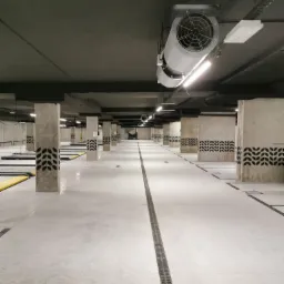 Wnętrze nowocześnie wykończonego parkingu podziemnego z betonowymi kolumnami ozdobionymi czarnym wzorem roślinnym, jasną posadzką z liniowym odpływem wody i dużym wentylatorem pod sufitem.