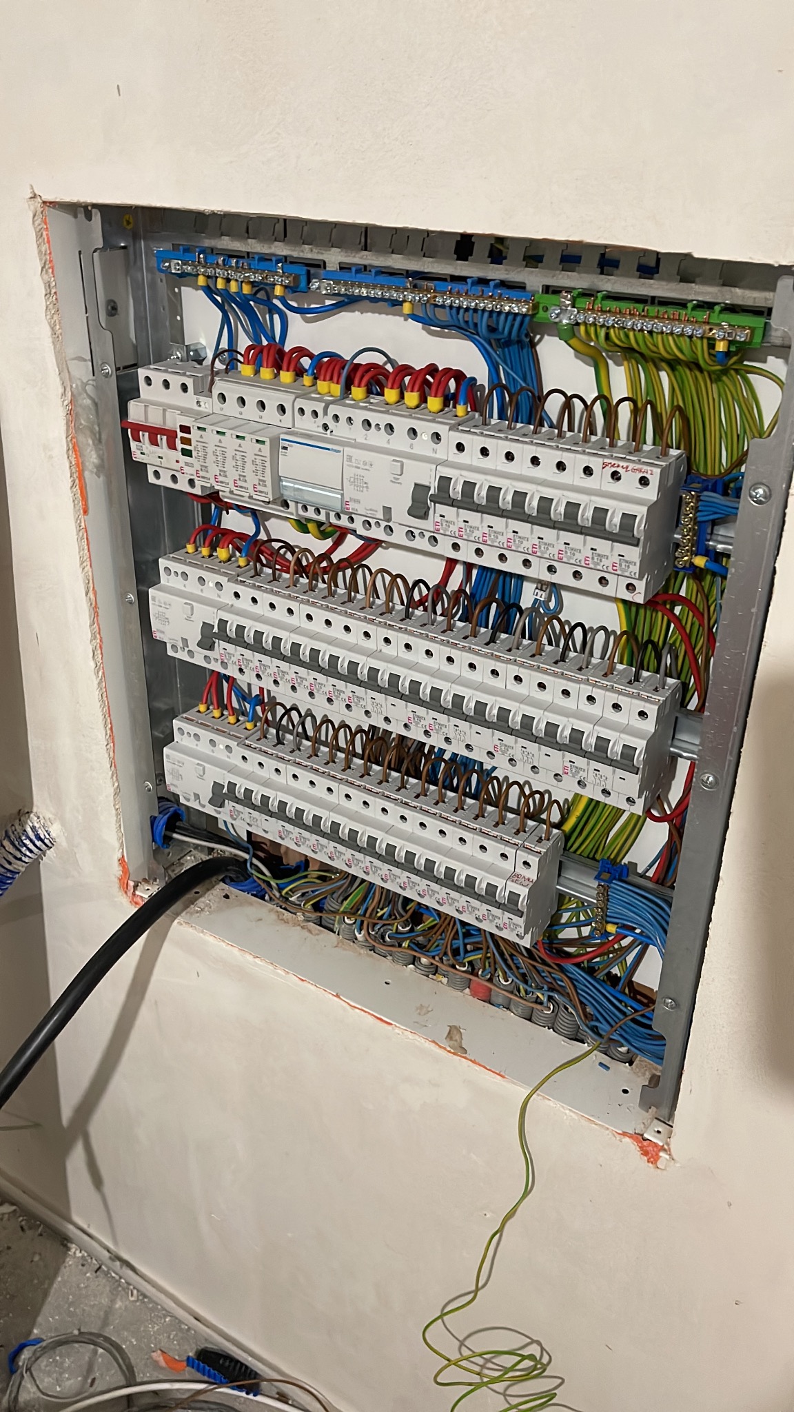 Rozdzielnica elektryczna z widocznymi przewodami w kolorach: niebieskim, żółto-zielonym, brązowym i czerwonym, umieszczona w ścianie, z widocznymi śladami prac wykończeniowych.