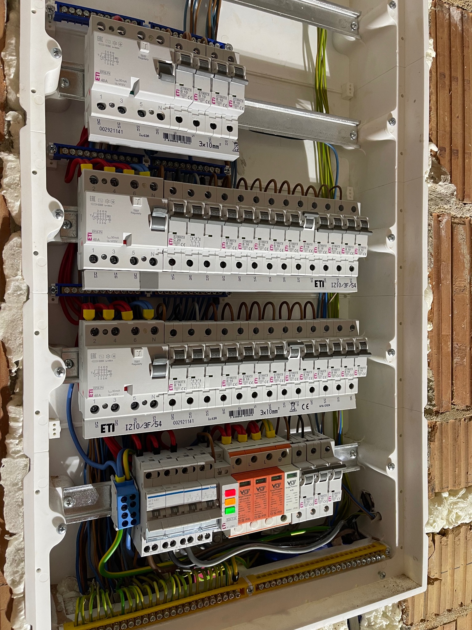 Rozdzielnica elektryczna z zamontowanymi bezpiecznikami ETI i VCX, widoczne przewody i listwa zaciskowa, instalacja w budynku z cegły.