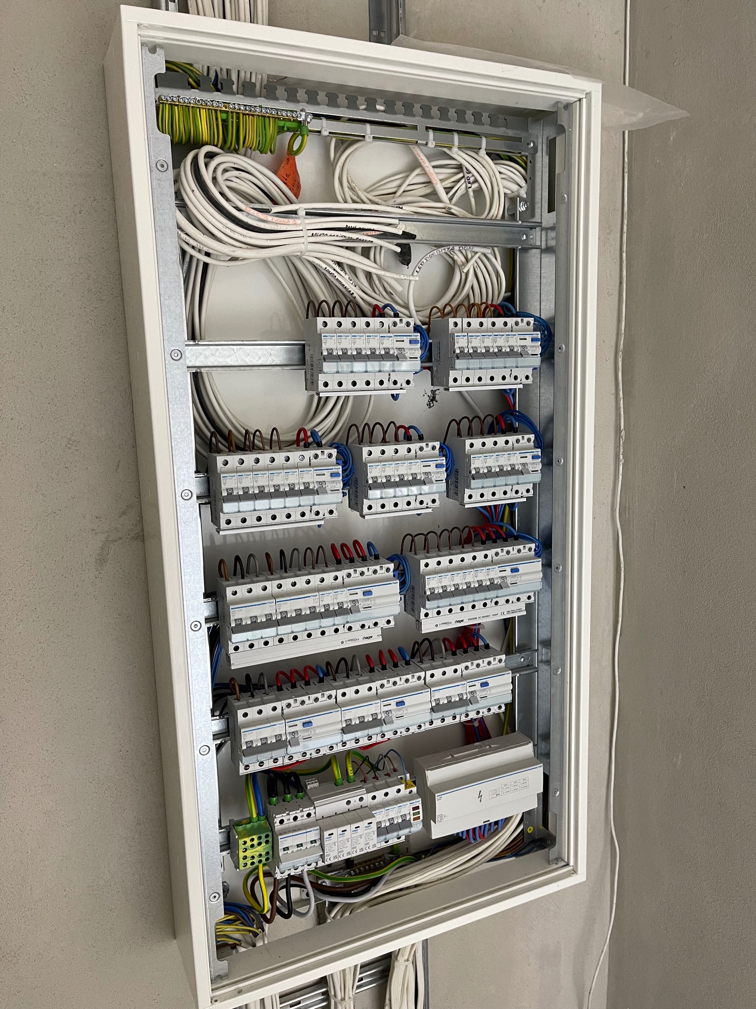 Otwarta skrzynka elektryczna z widocznymi bezpiecznikami, przewodami i listwami zaciskowymi, zamontowana na szarej ścianie. Widoczne kable uporządkowane wewnątrz skrzynki.