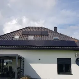 Dom jednorodzinny z ciemną dachówką i panelami fotowoltaicznymi, widoczne okna dachowe, pergola przy tarasie i antena satelitarna na elewacji.