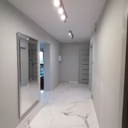 Wykończony korytarz w odcieniach szarości z marmurową podłogą, oświetleniem punktowym i dużym lustrem w ramie, widok na dalsze pomieszczenia.