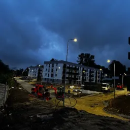 Oświetlona ulica w nowym osiedlu. Widoczne latarnie uliczne, budynek w budowie i maszyny budowlane na tle ciemnego nieba.