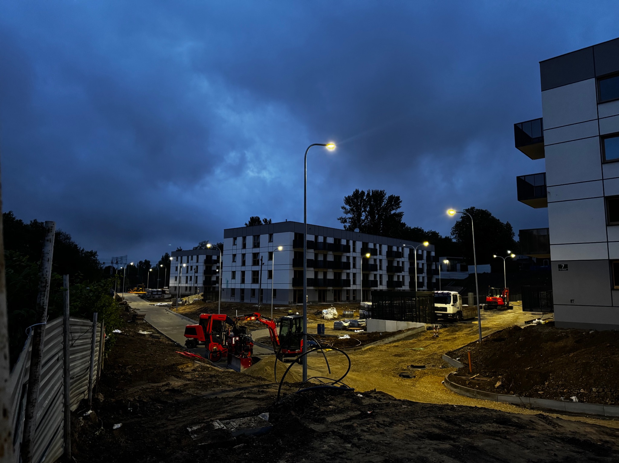 Nocny widok na plac budowy z oświetlonymi budynkami mieszkalnymi, maszynami budowlanymi i lampami ulicznymi pod ciemnym, zachmurzonym niebem.