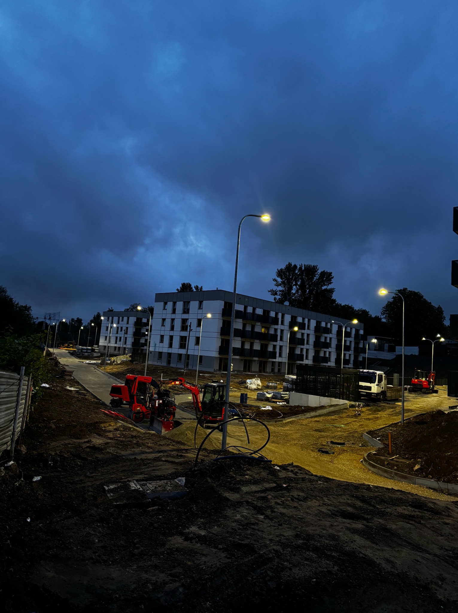 Oświetlona ulica w nowym osiedlu. Widoczne latarnie uliczne, budynek w budowie i maszyny budowlane na tle ciemnego nieba.