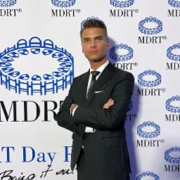 Mężczyzna w garniturze stojący na tle z powtarzającym się logo MDRT, z rękami skrzyżowanymi na piersiach i zegarkiem na nadgarstku.