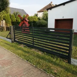 Nowoczesna, pozioma brama wjazdowa z metalowych profili w kolorze grafitowym, z widocznym czerwonym wiatrakiem w tle, oddzielająca posesję od chodnika z kostki.