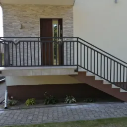 Zewnętrzne schody z czarną metalową balustradą i srebrnymi kulami dekoracyjnymi, prowadzące do drzwi wejściowych budynku z elewacją z kamienia dekoracyjnego.