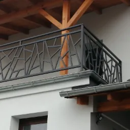 Nowoczesna balustrada balkonowa z czarnych metalowych profili o geometrycznym wzorze na tle białej ściany budynku z drewnianymi elementami konstrukcyjnymi.