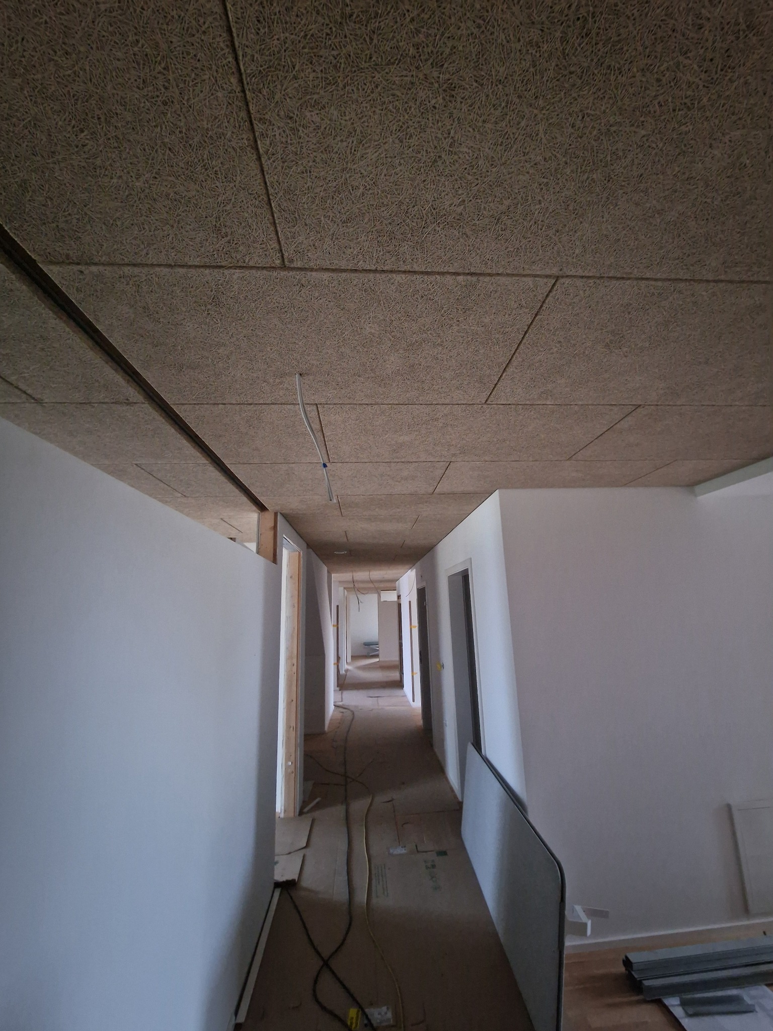 Korytarz w trakcie wykańczania: białe ściany, sufit z paneli akustycznych o fakturze drewna, widoczne kable i materiały budowlane. Ujęcie z perspektywy podłogi.