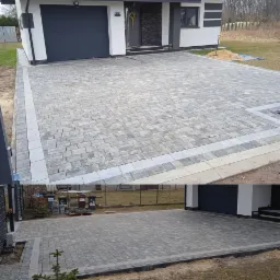 Ułożona kostka brukowa w odcieniach szarości na podjeździe przed nowoczesnym domem z garażem, widoczna sterta piasku z łopatą, obrzeża z jaśniejszej kostki.