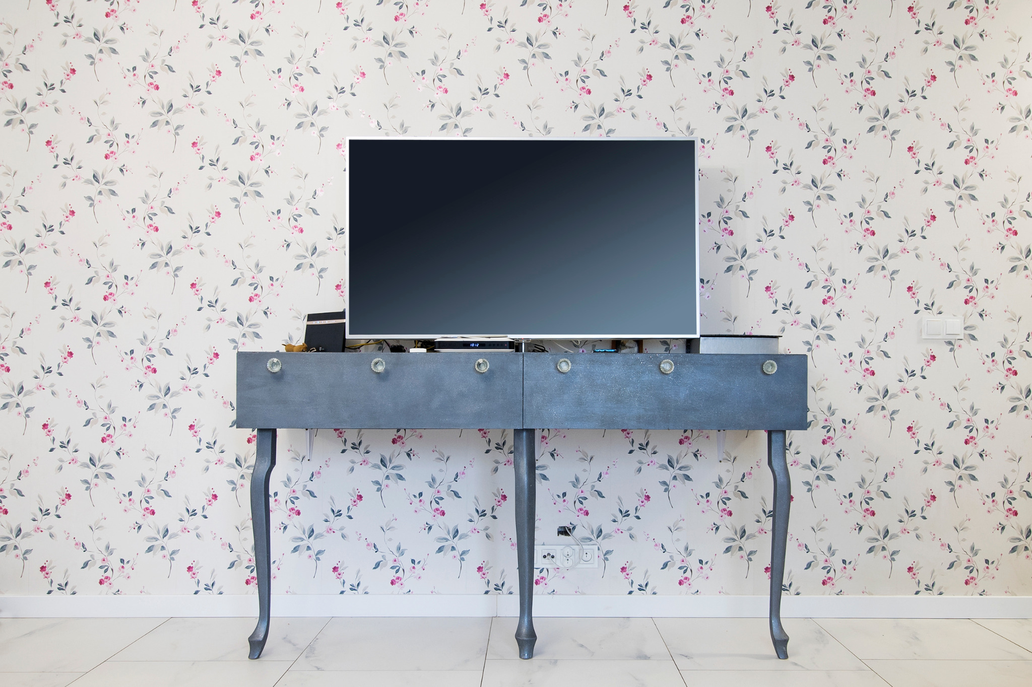 Telewizor z czarnym ekranem umieszczony na szarym, stylizowanym stoliku z kryształowymi gałkami na tle ściany w kwiatowy wzór.