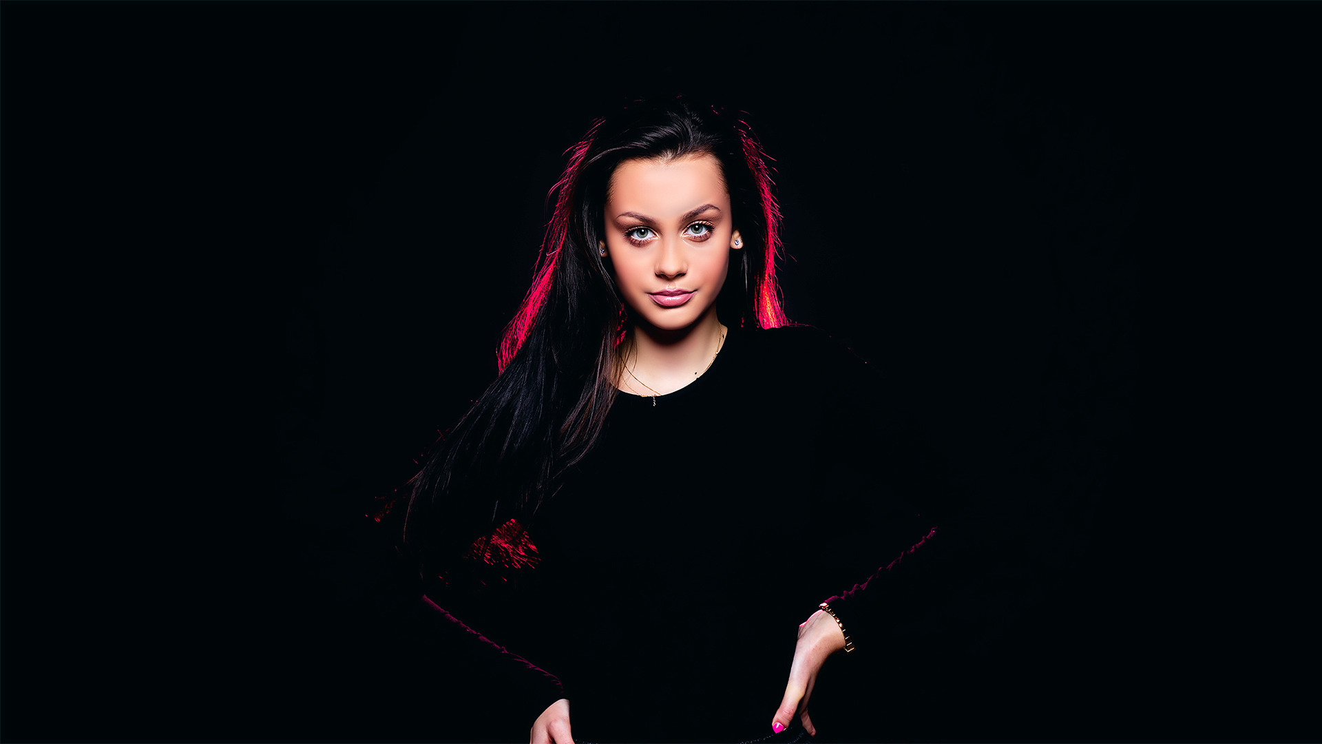 Portret młodej kobiety z długimi, ciemnymi włosami, oświetlonej czerwonym światłem od tyłu, na czarnym tle. Ubrana w czarny sweter, z delikatną biżuterią i subtelnym makijażem.