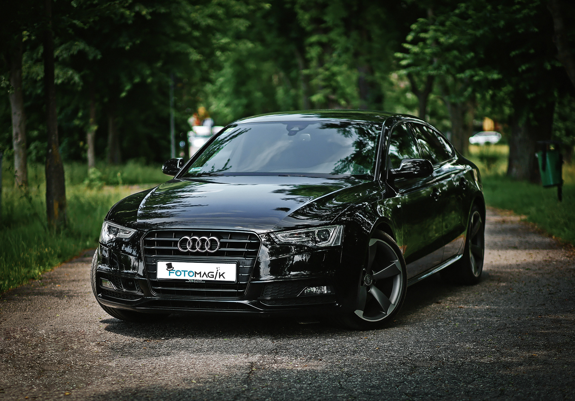 Czarny samochód Audi A5 na leśnej drodze, z logo Fotomagik na tablicy rejestracyjnej.