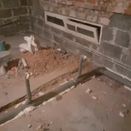 Szare rury kanalizacyjne ułożone w wykopie w surowym wnętrzu piwnicy, zabezpieczone pianką montażową, z widocznym oknem piwnicznym i gruzem.