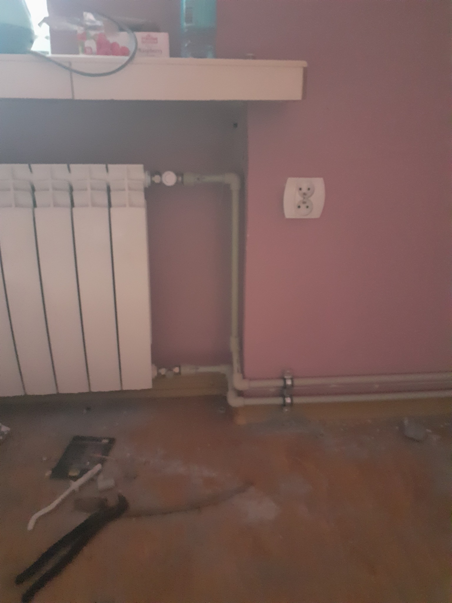 Biały grzejnik panelowy zamontowany na tle różowej ściany, z widoczną instalacją rur i narzędziami na podłodze