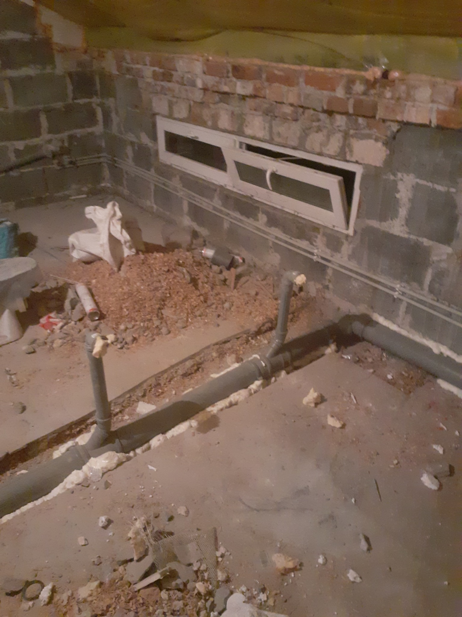 Szare rury kanalizacyjne ułożone w wykopie w surowym wnętrzu piwnicy, zabezpieczone pianką montażową, z widocznym oknem piwnicznym i gruzem.