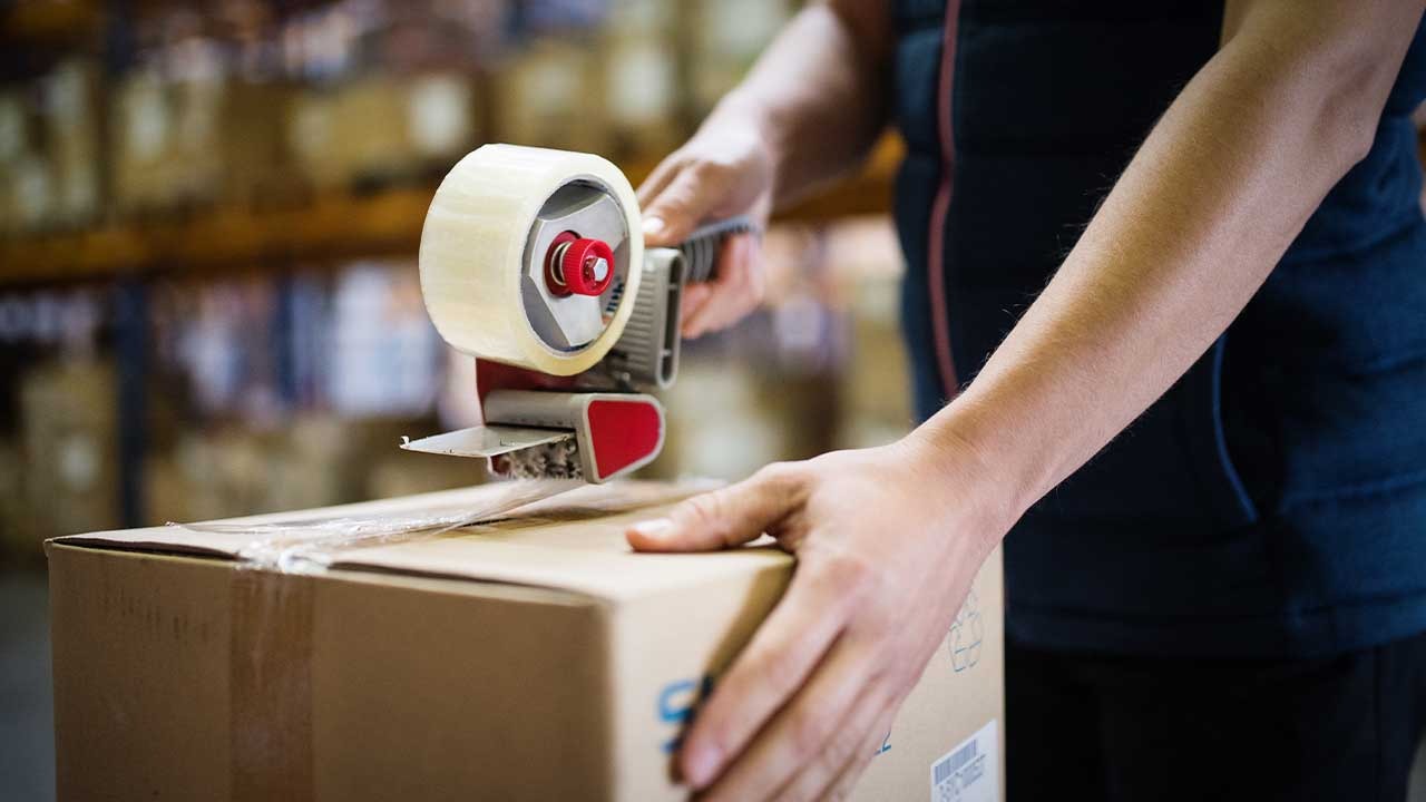 Osoba zakleja kartonowe pudełko dyspenserem z taśmą klejącą w magazynie, widoczne regały z kartonami w tle.