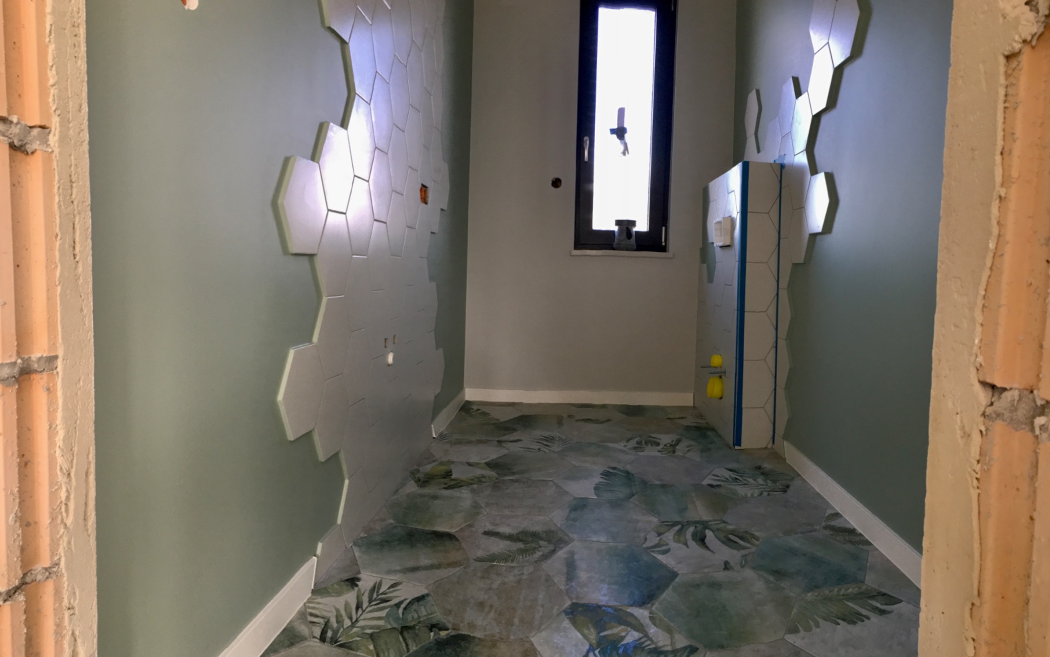 Niedokończona łazienka z heksagonalnymi płytkami na ścianie i podłodze, widoczny fragment surowej ściany z cegieł i okno z czarną ramą.