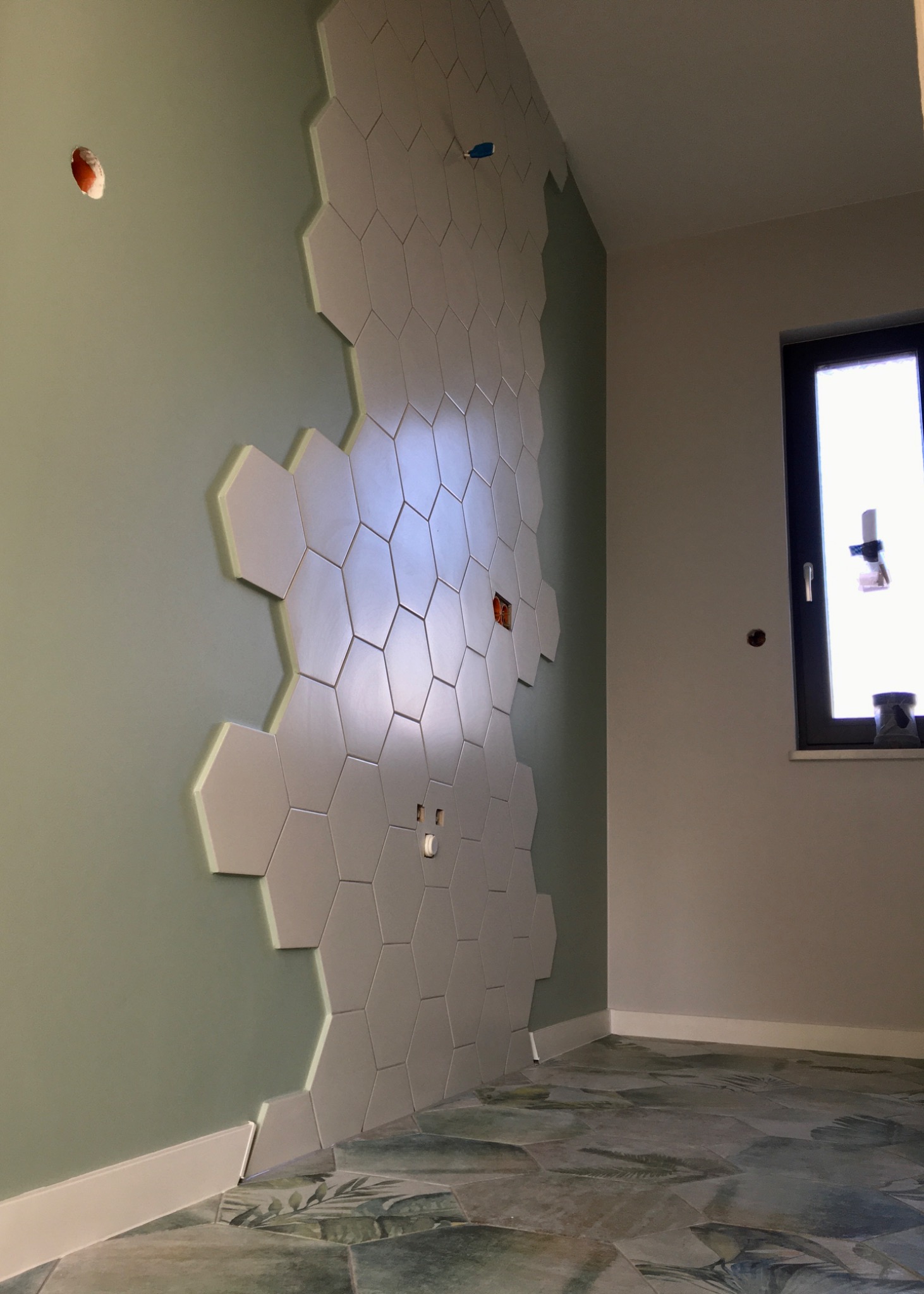 Ściana w trakcie wykańczania z częściowo ułożonymi heksagonalnymi płytkami ceramicznymi w odcieniach szarości, z widocznymi otworami na gniazdka elektryczne i fragmentem zielonej ściany z listwą...