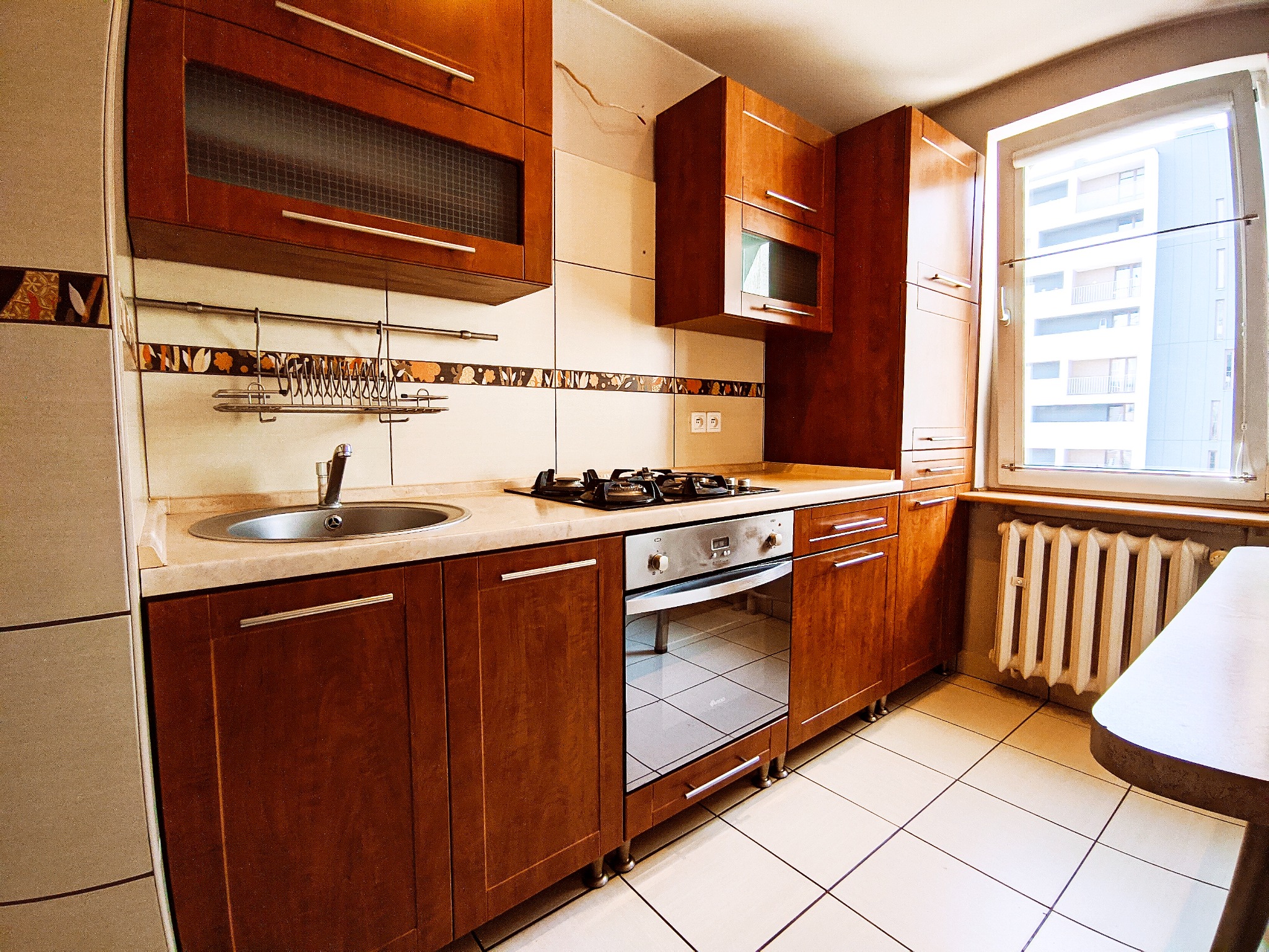 Ciemnobrązowa zabudowa kuchenna z beżowym blatem, widok na zlew, płytę gazową i piekarnik, białe płytki na podłodze i ścianie z dekoracyjnym pasem, okno z widokiem na budynek.