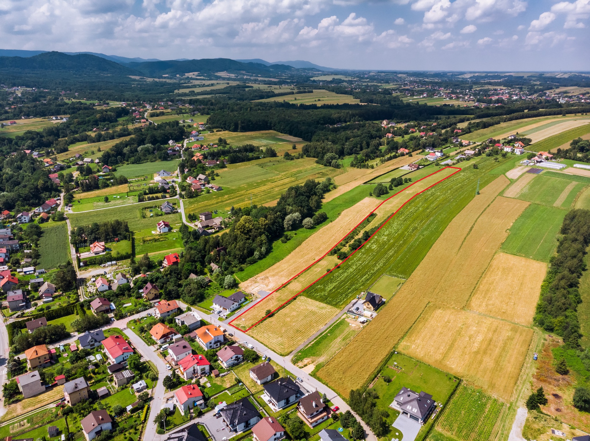 Widok z lotu ptaka na działkę budowlaną z wyraźnie oznaczonymi granicami, otoczoną polami uprawnymi i zabudową jednorodzinną, w tle zalesione wzgórza pod błękitnym niebem z chmurami.