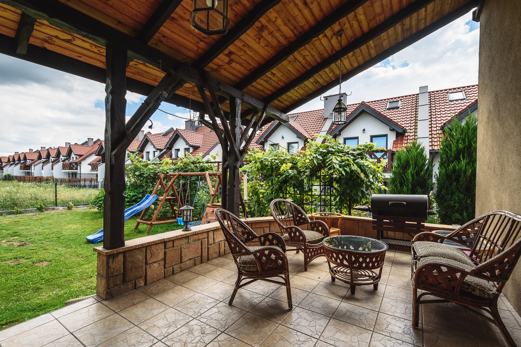 Zadaszony taras z rattanowymi meblami i grillem, widok na szereg domów z czerwonymi dachami i plac zabaw.