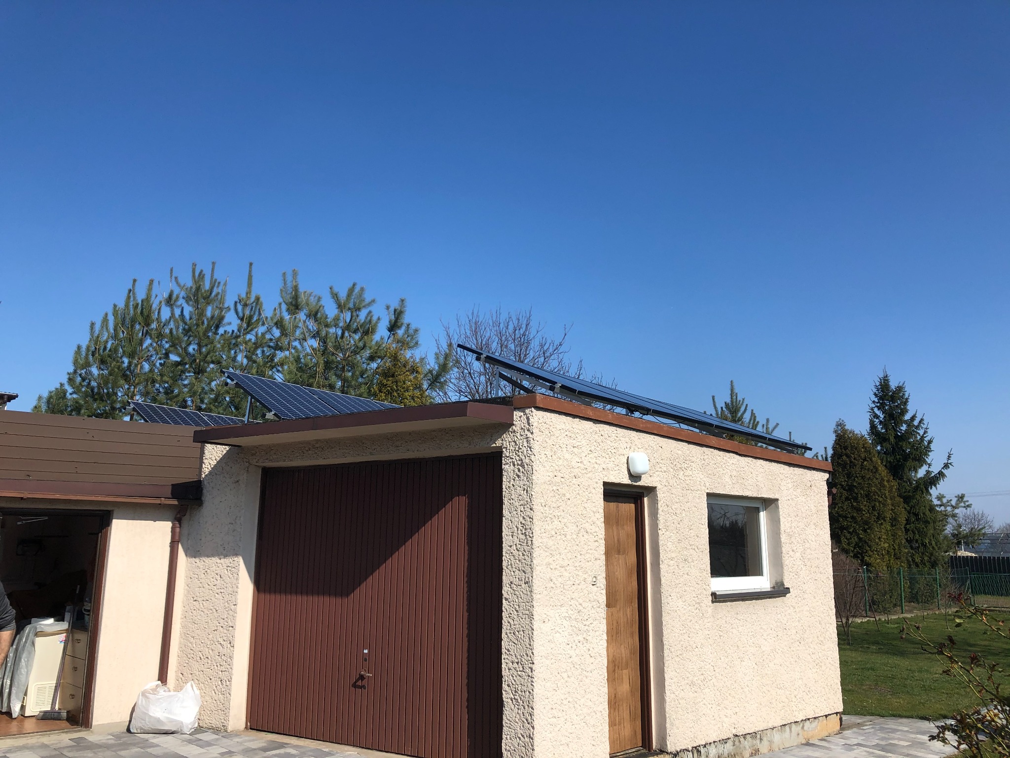 Panele słoneczne zamontowane na dachu garażu z brązową bramą i kremową elewacją w Gliwicach, widoczne drzewa iglaste i błękitne niebo.