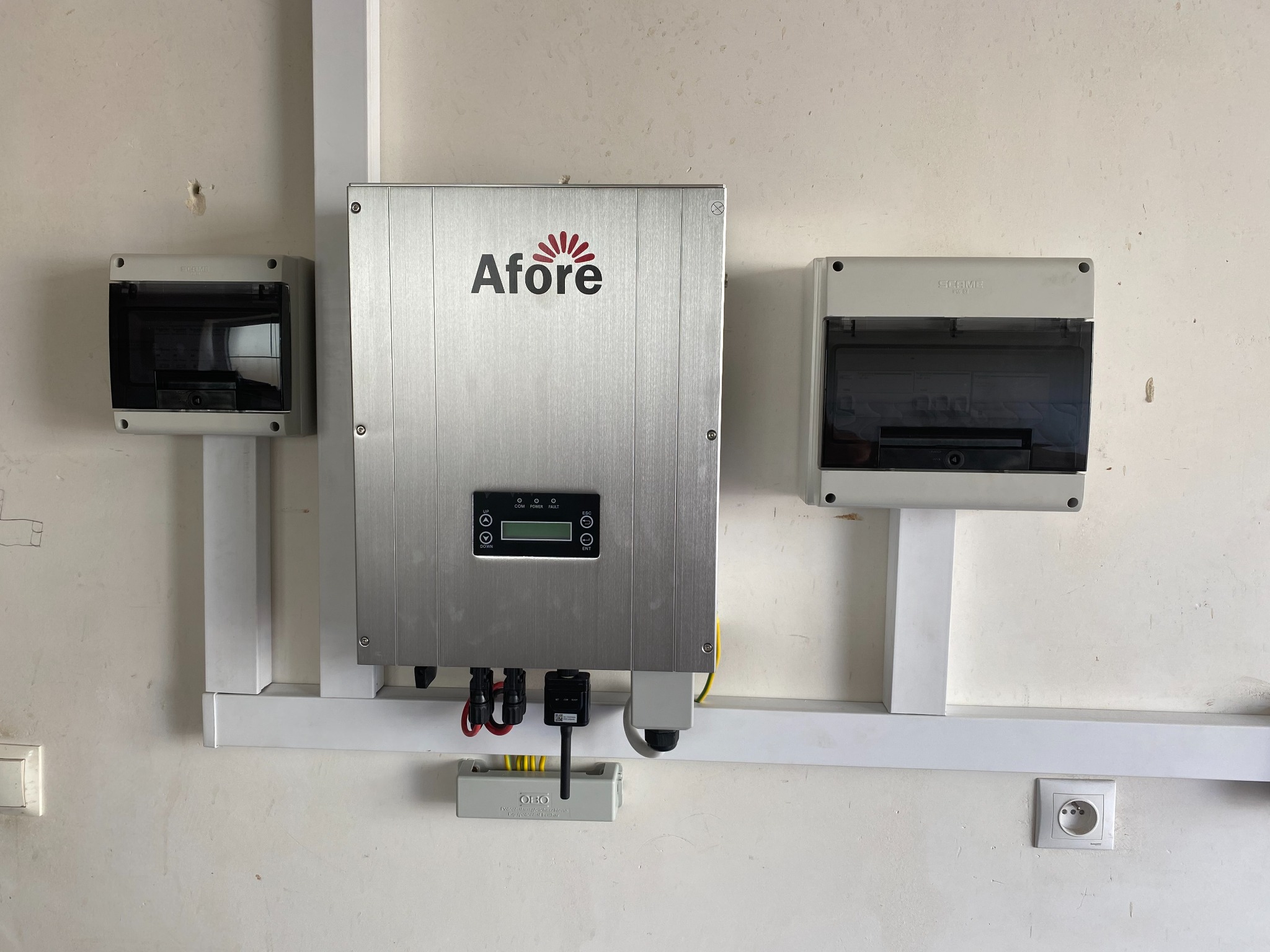 Zamontowany srebrny falownik Afore z podłączonymi kablami i zabezpieczeniami, widoczne skrzynki elektryczne po bokach, białe korytko kablowe i gniazdko elektryczne na tle ściany.
