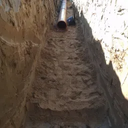 Wykop z widoczną rurą kanalizacyjną z tworzywa sztucznego oraz wiaderkiem, przygotowany do zasypania.