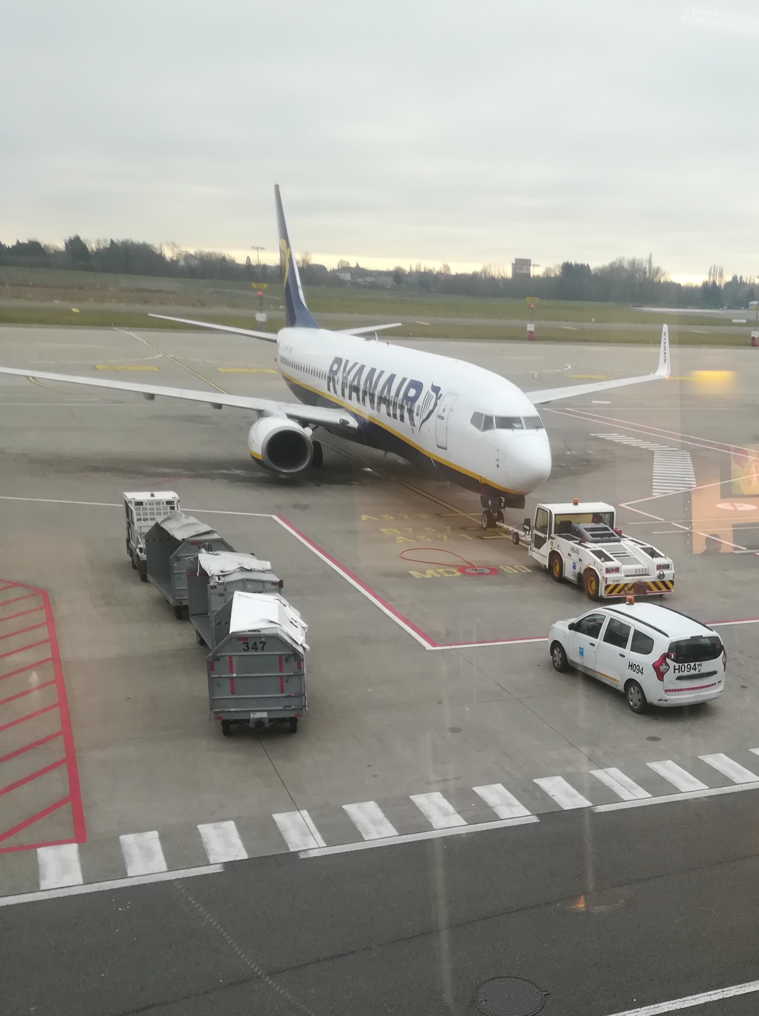 Samolot Ryanair na płycie lotniska, podłączony do holownika, obok wózek z bagażami i biały samochód obsługi lotniska z numerem H094.