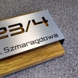 Metalowa tabliczka adresowa z wyciętym numerem 23/4 i nazwą ulicy 'ul. Szmaragdowa', zamontowana na drewnianej podstawie.