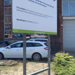 Tablica informacyjna o termomodernizacji budynków przemysłowych przy ulicy Fabrycznej 10 we Wrocławiu, z logo Funduszy Europejskich i Rzeczypospolitej Polskiej, na tle ceglanego budynku...