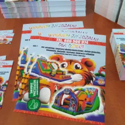 Stosy świeżo wydrukowanych ulotek i wizytówek z grafiką dmuchanych zamków i napisem 'Wynajem zjeżdżalni dla dzieci', leżące na drewnianym blacie.