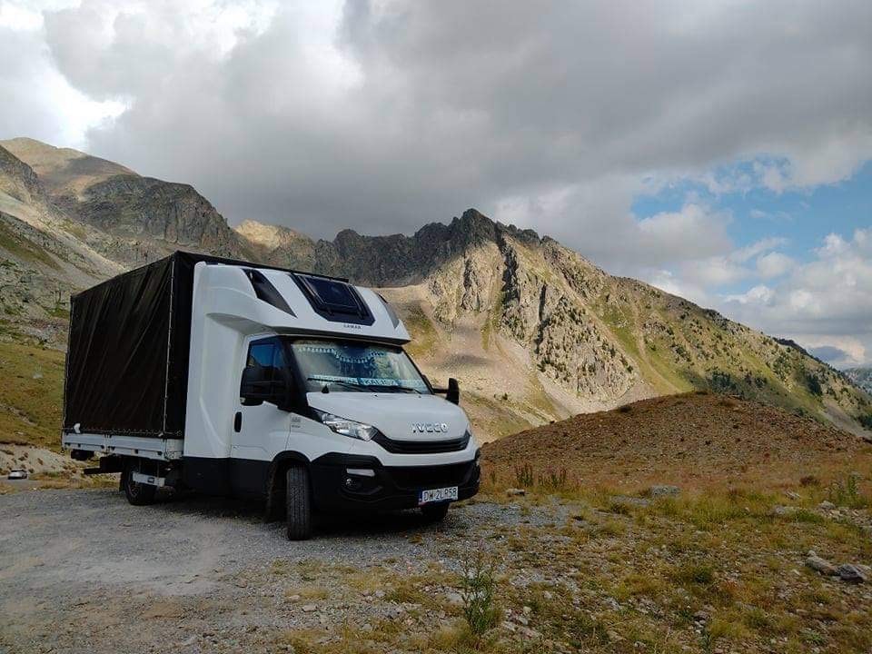 Biała ciężarówka Iveco z czarną plandeką zaparkowana na tle górskiego krajobrazu z pochmurnym niebem.