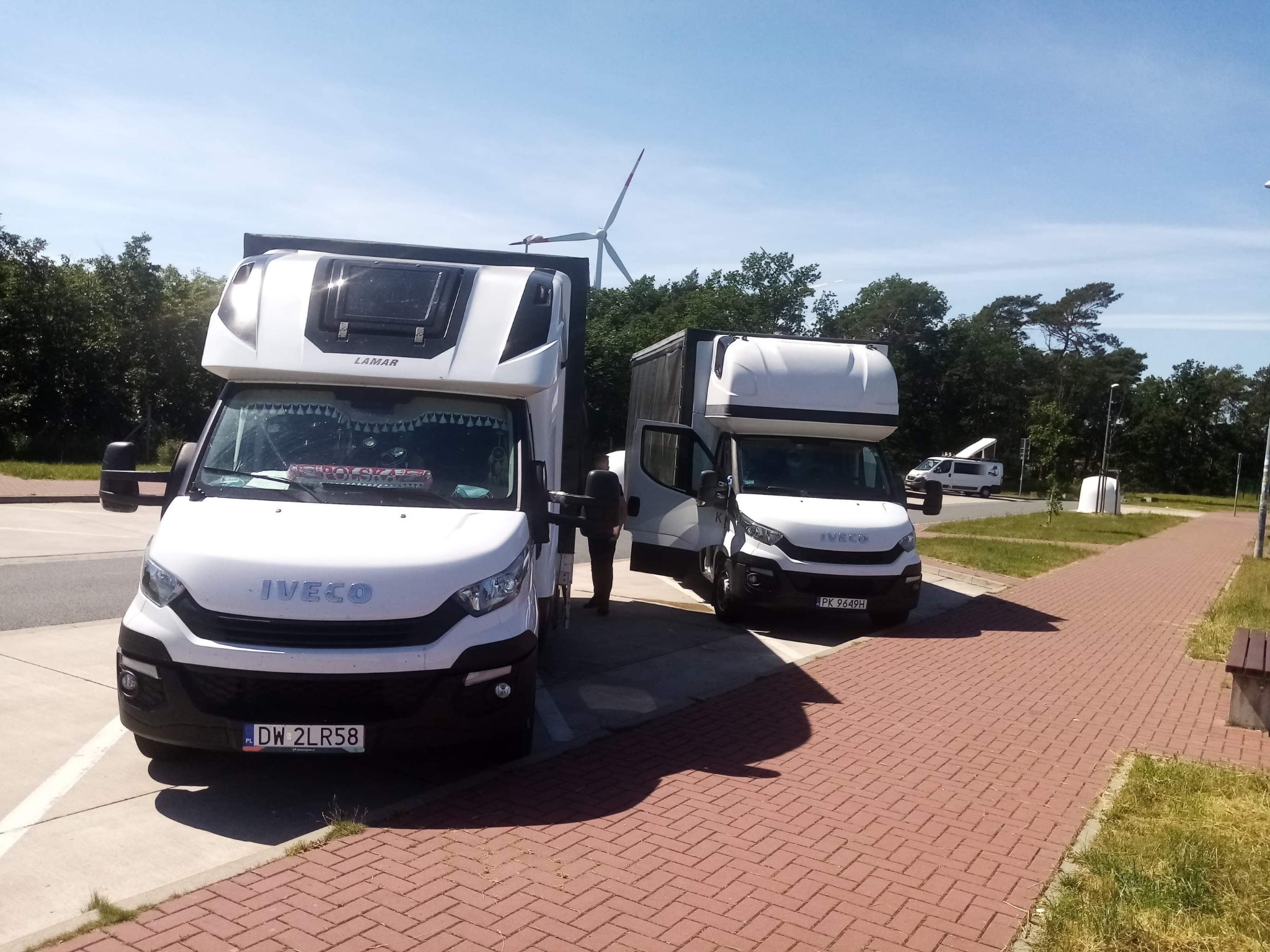 Dwa białe busy Iveco z nadbudową typu kontener, zaparkowane obok siebie na parkingu z kostki brukowej, w tle widoczny wiatrak i zieleń, jeden z busów ma otwarte drzwi kierowcy.