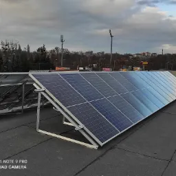 Rząd paneli fotowoltaicznych zamontowany na płaskim dachu budynku komercyjnego, widoczne metalowe konstrukcje wsporcze, w tle zabudowa miejska i pochmurne niebo.