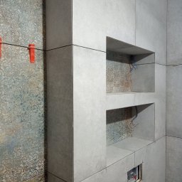 Kostrzewski - Wnętrze łazienki w trakcie wykańczania: szare płytki z dekoracyjnym wzorem, wnęki w ścianie i czerwone klipsy poziomujące płytki. Widoczne kable elektryczne.