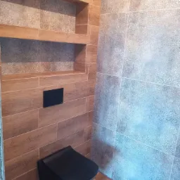 Wykończona łazienka z drewnopodobnymi kaflami, wnękami i szarą ścianą z delikatnym wzorem. Czarna toaleta podwieszana. Nowoczesny design z akcentem naturalnego drewna.