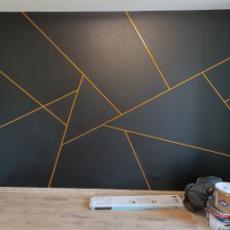 Kostrzewski - Czarna ściana z geometrycznym wzorem w odcieniach złota, świeżo po malowaniu. Na podłodze widoczne akcesoria malarskie i puszki po farbie. Nowoczesny design.