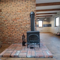 Kostrzewski - Rustykalny piec żeliwny z kominem na ceglanym podeście, na tle ceglanej ściany i drewnianego stropu. Widoczne akcesoria kominkowe i fragment wykończonej kuchni w tle.