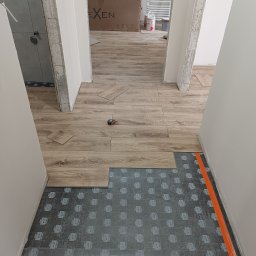 Kostrzewski - Układanie paneli podłogowych w korytarzu. Widoczna podkładka pod panele i częściowo ułożona podłoga z paneli imitujących drewno. Pomarańczowa taśma zabezpieczająca przy ścianie.