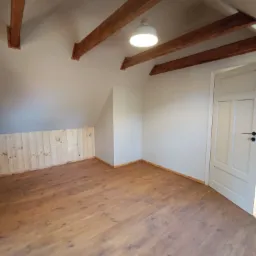 Jasne poddasze z drewnianą podłogą i boazerią. Widoczne drewniane belki stropowe i białe drzwi. Całość oświetlona centralnym plafonem.