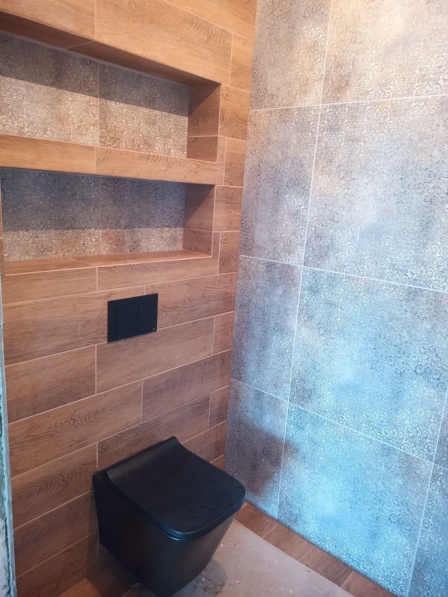 Wykończona łazienka z drewnopodobnymi kaflami, wnękami i szarą ścianą z delikatnym wzorem. Czarna toaleta podwieszana. Nowoczesny design z akcentem naturalnego drewna.