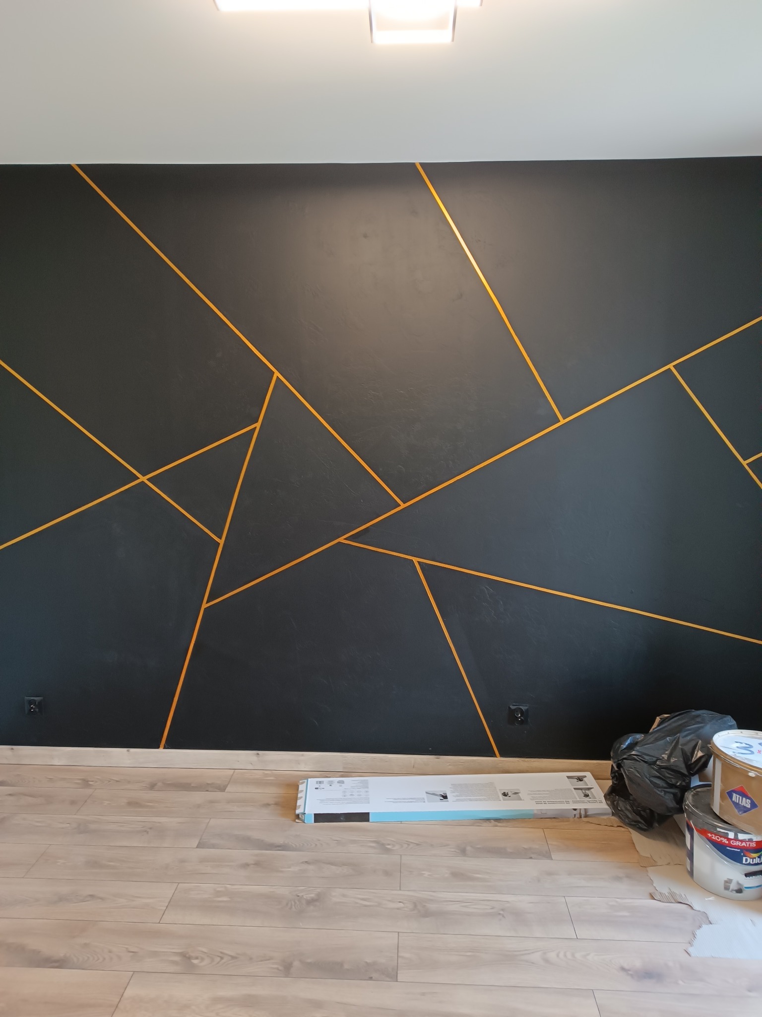 Czarna ściana z geometrycznym wzorem w odcieniach złota, świeżo po malowaniu. Na podłodze widoczne akcesoria malarskie i puszki po farbie. Nowoczesny design.