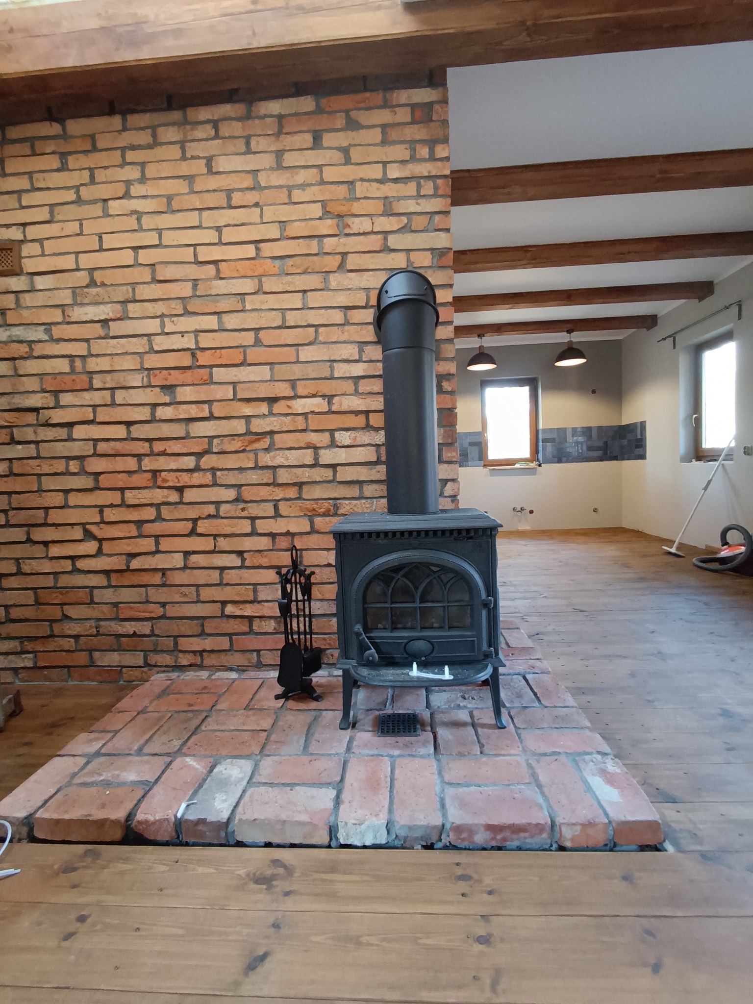 Rustykalny piec żeliwny z kominem na ceglanym podeście, na tle ceglanej ściany i drewnianego stropu. Widoczne akcesoria kominkowe i fragment wykończonej kuchni w tle.